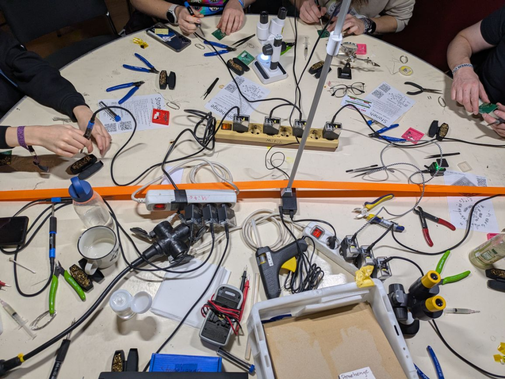 A somewhat messy table. lots of soldering irons, wires, tweezers, a microscope, instruction manuals and various other tools. 
The hands of 4 people working on the stonehenge board can be seen.